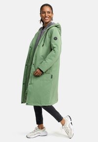 Long green outerwear coat with a hood, gray lining, and black accents. Features snap buttons and side pockets, styled with dark jeans and light sneakers.