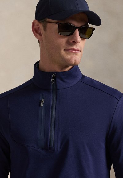 Μακρυμάνικο πουλόβερ σε ναυτικό μπλε με φερμουάρ, ψηλό γιακά, τσέπη στο στήθος με φερμουάρ και απαλό υλικό, συνδυασμένο με καπέλο και γυαλιά ηλίου στο ίδιο χρώμα.