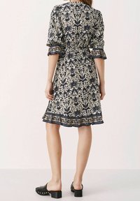 Robe à motif floral en crème et bleu marine, avec des manches trois-quarts, une taille ceinturée et un ourlet volanté. Portée avec des mules à talons noirs.
