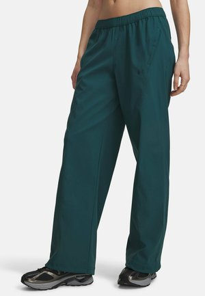 Lower torso of a person wearing loose dark green pants with elastic waist and black athletic shoes, standing against a plain background.