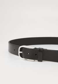 Curea din piele neagră cu cataramă rectangulară argintie, gravată cu "BOSS", întinsă pe o suprafață albă.