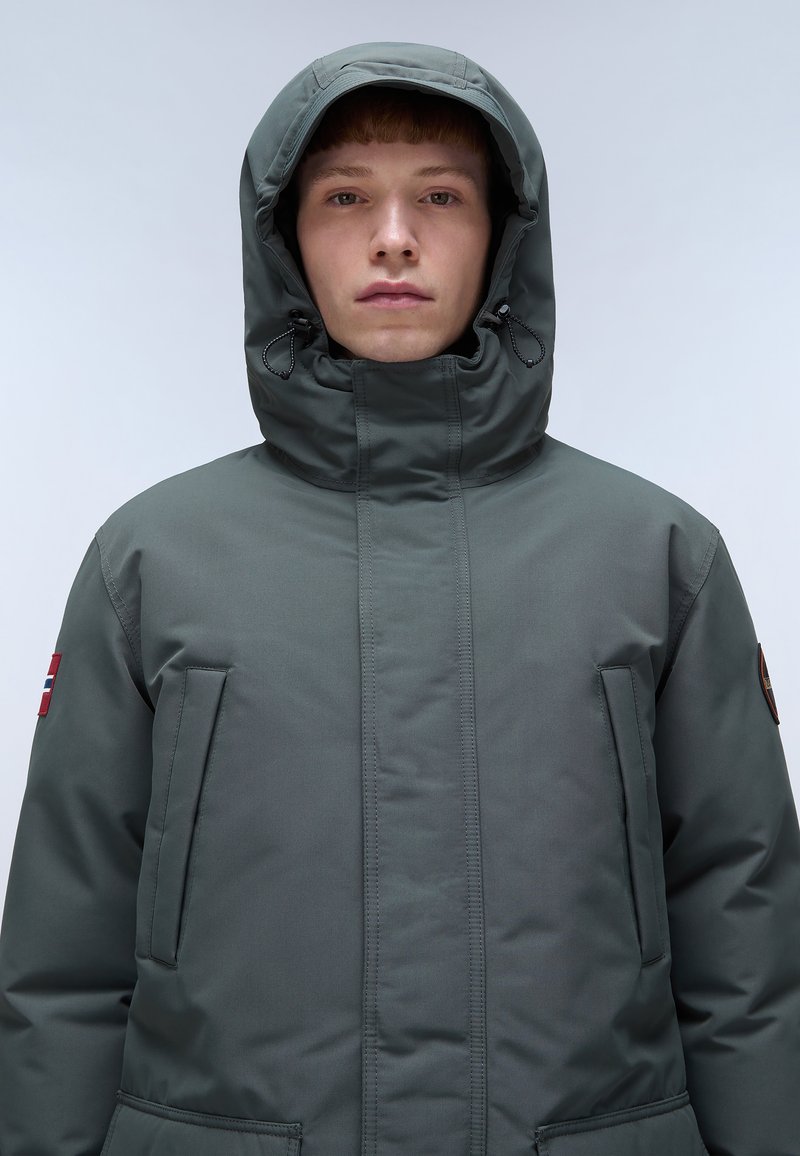 Veste doudoune verte avec un col montant et une capuche ajustable. Dotée de poches zippées et d'un patch sur la manche gauche. Texture de tissu lisse.