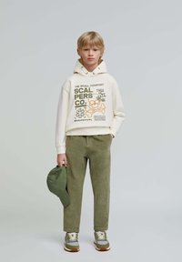 Felpa crema con stampe grafiche, pantaloni texturizzati verde oliva, cappellino verde in mano e sneakers grigie con lacci crema e suole marroni.