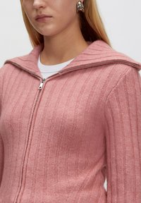 Nahaufnahme einer Person, die einen pinkfarbenen, gerippten Reißverschlusspullover über einem weißen Hemd trägt, ergänzt mit großen silbernen Creolen.