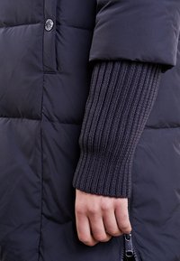 Hand i mörk ribbad stickad ärm bredvid en svart vadderad jacka med knapp och delvis neddragen dragkedja.