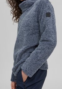 Meszysty niebieski polarowy polar z wysokim kołnierzem. Posiada małą naszywkę z logo na lewym rękawie oraz szerokie mankiety dla wygody.