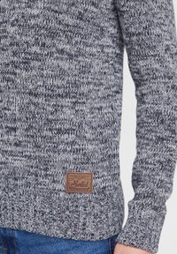 Strikket sweater med et struktureret gråt og marineblåt mønster. Inkluderer et brunt lædermærke med "Solid" branding forneden.