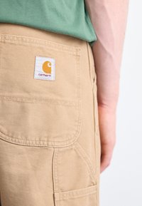 Tan Carhartt-broeken met een textuur, voorzien van een achterzak en een wit label met een oranje logo, zijaanzicht.