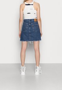 Woman wearing a white tie-dye tank top, blue denim skirt with frayed hem, and white high-top sneakers with black text on the back.