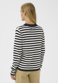 Donna con capelli biondi che indossa un maglione a righe orizzontali bianche e nere e pantaloni beige, girata di spalle contro uno sfondo semplice.