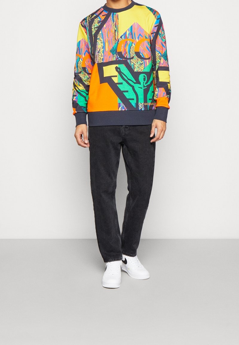 Sweat-shirt coloré présentant un mélange de motifs verts, orange, bleus et jaunes, avec un design graphique et des poignets côtelés, associé à un jean foncé.