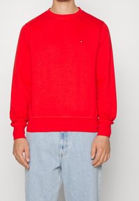 Rode katoenen sweatshirt met ronde hals, lange mouwen en geribbelde manchetten en zoom. Bevat een klein logo op de borst. Gedragen met lichtblauwe jeans.