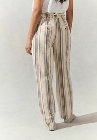 Pantalons larges rayés en crème avec des rayures verticales rouges, vertes et noires. Taille élastiquée, poches arrière et tissu texturé.