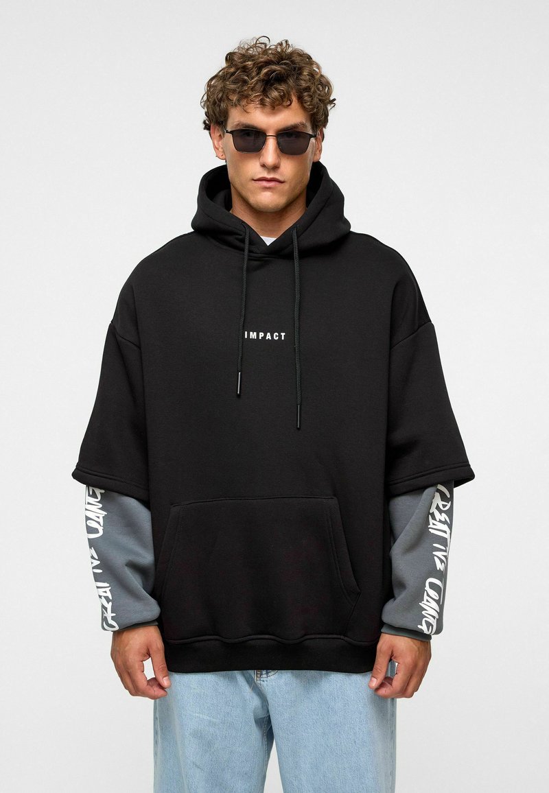 Zwarte oversized hoodie met grijze mouwaccenten, voorzien van een voorzak en "IMPACT" gedrukt op de borst. Gladde katoenmix stof.