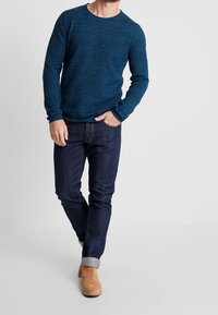 Pull en maille bleu foncé avec un col rond, associé à un jean foncé et des bottines camel. Le jean a un revers retroussé à la cheville.