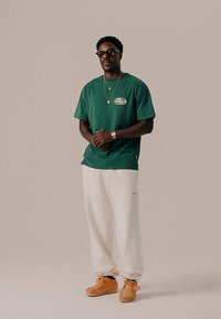 Muž v zeleném tričku, bílých teplákových kalhotách, béžových botách, slunečních brýlích a zlatých šperkách stojící s rukama spojenýma proti neutrálnímu pozadí.