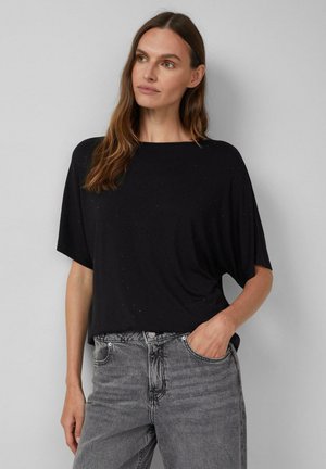 Vrouw met lang bruin haar, gekleed in een losse zwarte top met korte mouwen met subtiele stippen en hoog opgesneden grijze jeans, hand in zak, kijkt naar links.