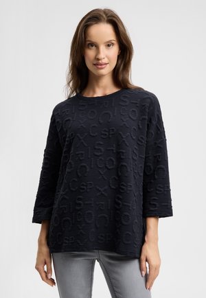 Marineblaue Sweatshirt mit strukturiertem Logo-Muster, rundem Ausschnitt und dreiviertel Ärmeln, kombiniert mit hellgrauen Jeans.