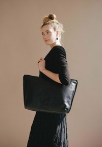Borsa tote nera in pelle con una forma rettangolare spaziosa e una texture liscia, tenuta da un modello che indossa un maglione scuro su uno sfondo neutro.
