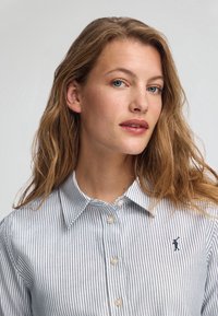Camicia button-up a righe azzurro chiaro e bianco con colletto classico, presenta piccoli bottoni rotondi e un ricamo del logo blu navy sul petto.