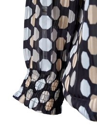 Blouse transparente avec un motif hexagonal noir, blanc et beige, ornée de poignets froncés et d'une texture légère et fluide.