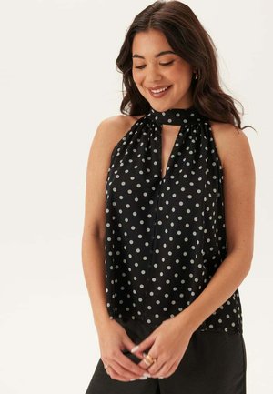 Kvinna som ler, klädd i ärmlös svart topp med vita prickar och hög krage med nyckelhålsutskärning, händerna sammanfogade framför sig.