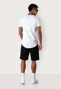 White short-sleeve t-shirt with mint green vertical text on the back. Paired with black shorts and white sneakers. Smooth fabric, round hem.