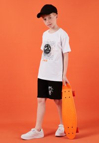 Garçon portant une casquette noire, un t-shirt blanc à thème spatial, un short noir avec un imprimé d'astronaute, des baskets blanches, tenant un skateboard orange sur un fond orange.