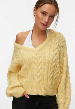 Junge Frau trägt einen locker sitzenden gelben Strickpullover mit Zopfmuster, der von einer Schulter rutscht, darunter ein weißes Neckholder-Top, und schwarze Hosen, posiert mit der Hand in der Tasche.