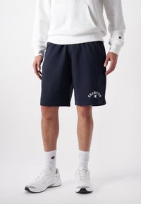 Navy cotton shorts with an elastic waistband and side pockets, featuring a white "Champion" logo in a curved style. Paired with white sneakers.