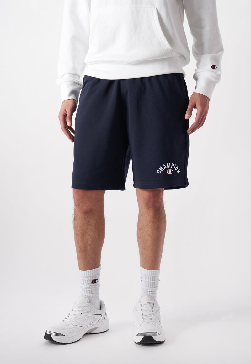 Marineblauwe katoenen korte broek met een elastische tailleband en zijzakken, voorzien van een witte "Champion"-logo in een gebogen stijl. Gekoppeld aan witte sneakers.