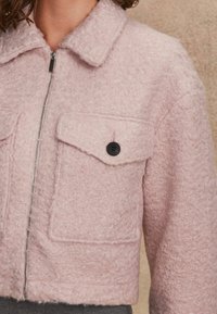 Růžová, strukturovaná zkrácená bunda vyrobená z měkkého materiálu, se zapínáním na zip, dvěma náprsními kapsami s klopami a knoflíkovým uzávěrem a límcem.