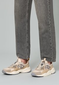Sneakers beige e grigi con un mix di materiali scamosciati e in rete, silhouette robusta, suola testurizzata e dettagli dei lacci.
