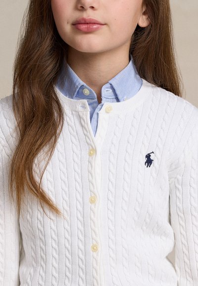 Jeune personne aux longs cheveux bruns portant un cardigan en maille torsadée blanche avec des boutons sur une chemise bleu clair à col.