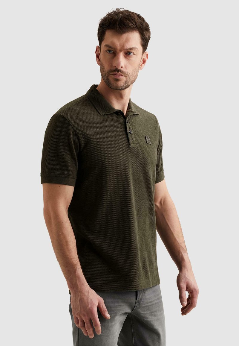 Homme aux cheveux courts et foncés portant un polo vert olive texturé et un jean gris, debout devant un fond clair uni.