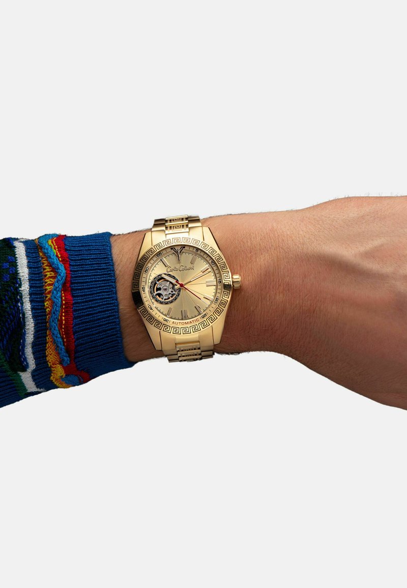 Gold-Armbanduhr mit geometrischem Muster auf der Lünette, rundem Zifferblatt, silbernen Akzenten und automatischem Uhrwerk, das durch einen transparenten Bereich sichtbar ist.