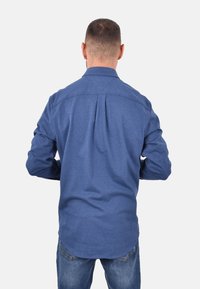 Camicia blu a maniche lunghe realizzata in tessuto morbido, con yoke posteriore, orlo curvo e colletto button-down, abbinata a jeans in denim.