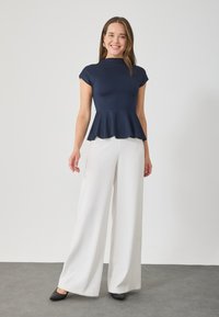 Top peplum navy con mangas cortas combinado con pantalones anchos blancos y tacones negros, de pie frente a un fondo neutro.
