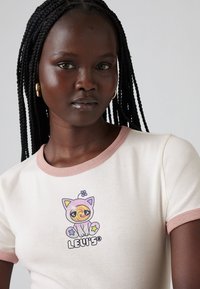 Ung kvinne med langt fletthår har på seg en lys beige t-skjorte med rosa detaljer og en fargerik tegneserie-kattgrafikk på forsiden.