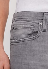 Szare dżinsy charakteryzują się slim fit, kontrastowym szyciem, małą przednią kieszenią oraz metalowymi nitami w pasie dla dodatkowego akcentu.