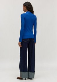 Blaues gestreiftes Langarmoberteil mit hohem Kragen, kombiniert mit weit geschnittenen dunklen Jeans mit hochgerollten Säumen und braunen High Heels.