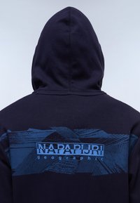 Sweat à capuche marine avec impression graphique bleue mettant en avant le texte "NAPAPIJRI geographic". Design texturé avec une capuche proéminente et des détails de couture.