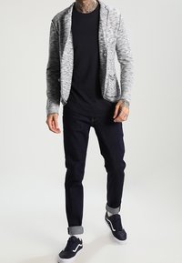 Blazer gris jaspeado sobre una camisa negra, combinado con jeans oscuros enrollados y zapatillas azul marino. Presenta un cuello de chal y bolsillos frontales.