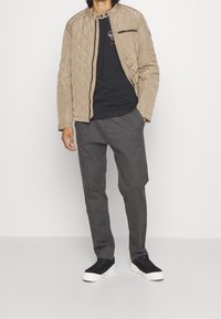 Světle béžová prošívaná bunda s vystouplým límcem, černou kapsou na zip, v kombinaci s tmavě šedými kalhotami a černými teniskami. Jednotná textura a design.