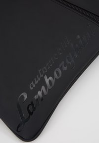 Černá peněženka s hladkým povrchem, na přední straně s emblémem "automobili Lamborghini" a se zipovým uzávěrem podél horního okraje.