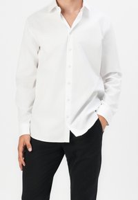 Weißes Hemd mit langen Ärmeln und Kragen, aus glattem Stoff, kombiniert mit schwarzen Hosen, die einen lässigen Schnitt haben.