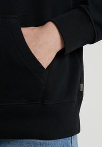Felpa nera con tasca a marsupio, polsini a coste e una piccola etichetta metallica sul lato. Tessuto morbido con una texture liscia.