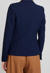 Chaqueta ajustada de color azul marino con tejido texturizado, con una abertura en la parte trasera y puños con botones, llevada sobre pantalones marrones, mostrada desde la parte de atrás.
