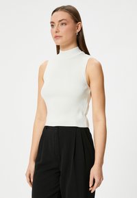 Witte mouwloze turtleneck trui met ribbelstructuur, slim fit en contrasterende zijpanelen, gecombineerd met zwarte geplooide broeken.