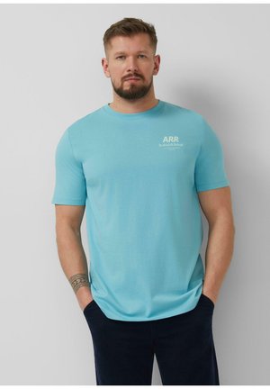 Mann mit Bart, der ein hellblaues ARR Rethink & Reload T-Shirt und schwarze Hose trägt, vor einem schlichten hellgrauen Hintergrund stehend.